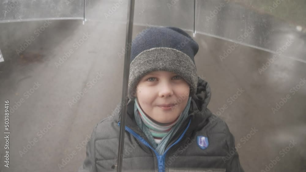 Cute caucasian boy in warm jacket and hat looks and smiles through wet transparent dome of umbrella with water drops. Rainy weather and wet asphalt with puddles. Cute caucasian boy in warm jacket and hat looks and smiles through wet transparent dome of umbrella with water drops. Rainy weather and wet asphalt with puddles.