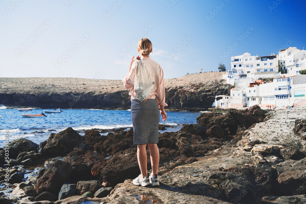 Stylish woman with fruits in cotton bag at beach, Canary Islands, Spain. Happy young girl tourist relaxing, enjoying picnic in small village on vacation. Concepts low waste, eco-travel, local tourism.