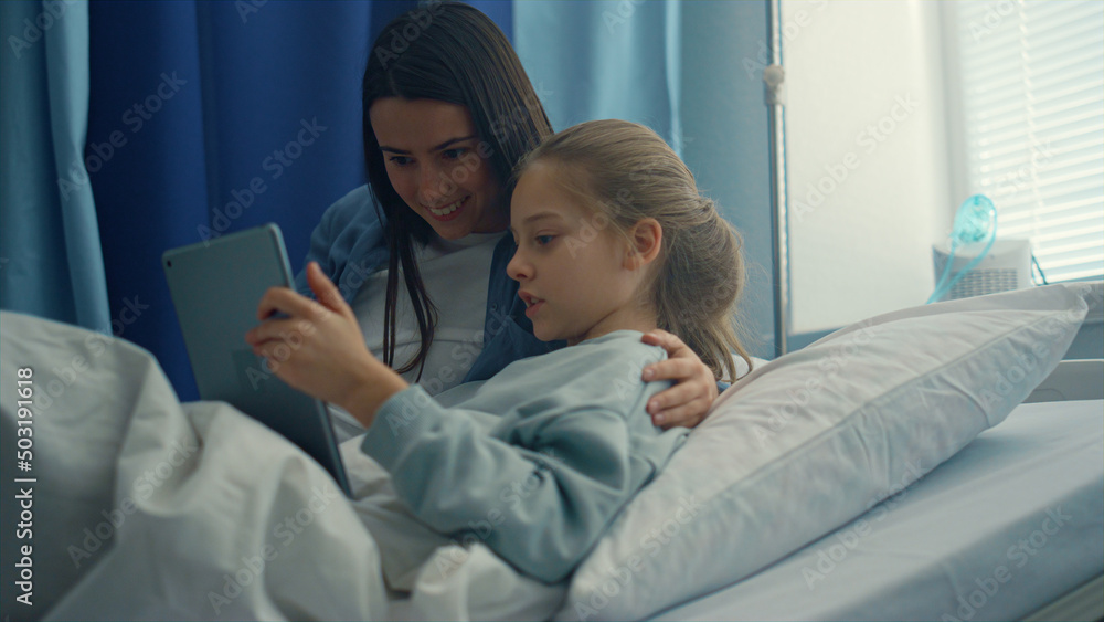 Smiling mom visiting daughter play on tablet computer together in hospital room