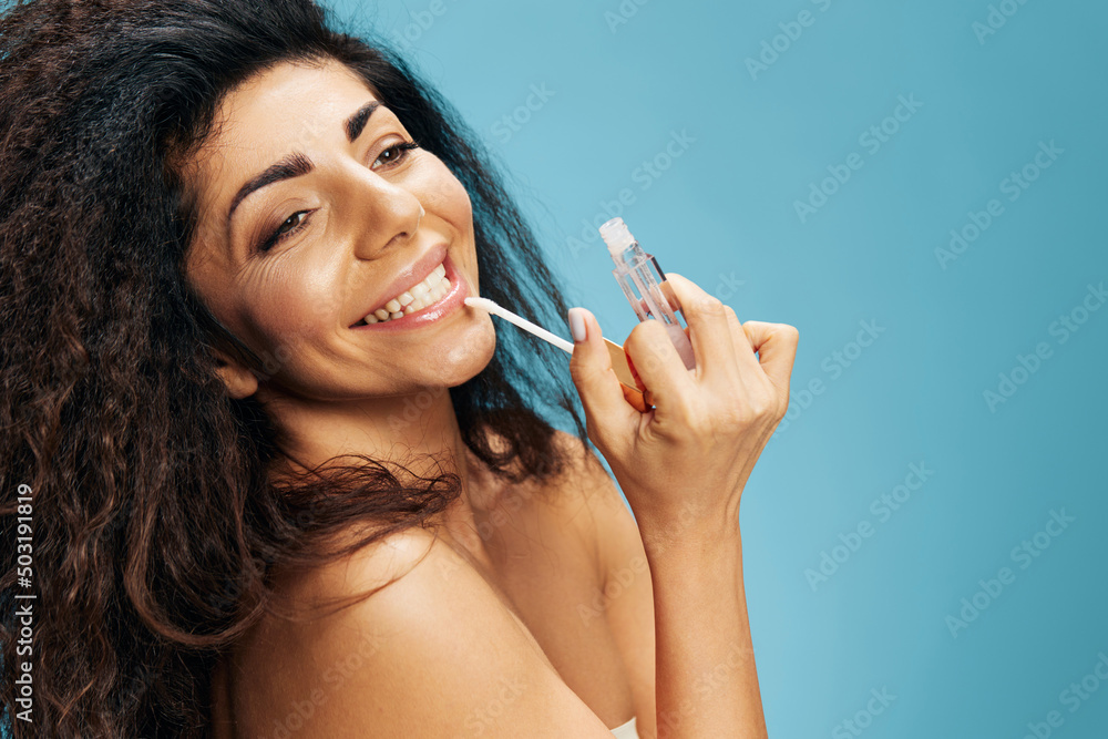 Obraz premium Beautiful smiling young tanned curly latin woman look aside paint lips applying tender moisturizing lipstick do visage facial makeup using lip gloss isolated blue background. Copy space