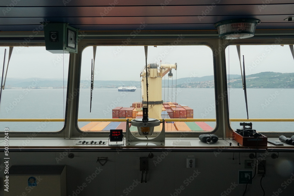 Koper, Slovenia - 05 06 2022: View through central window with gyro repeater of navigational bridge of container vessel to the ro-ro ship leaving the port of Koper, Slovenia during rainy day.