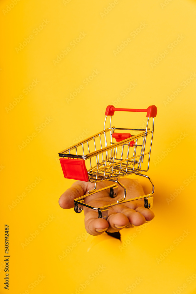 Male hand holds through a hole a mini grocery shopping trolley on a yellow paper background close-up. Vertical image Male hand holds through a hole a mini grocery shopping trolley on a yellow paper background close-up. Vertical image