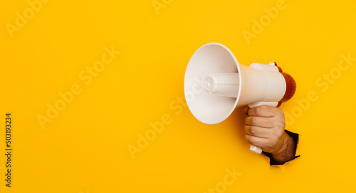 Fototapeta Naklejka Na Ścianę i Meble -  Hand holds a megaphone from a hole in the wall on a yellow background with copy space. Concept of hiring, advertising something