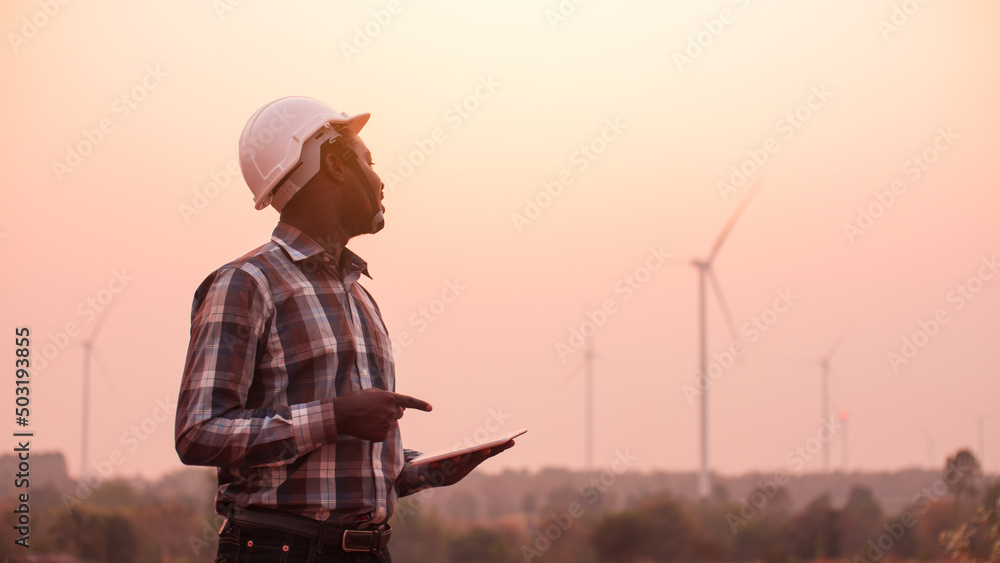 African engineer man stands holding tablet front the wind turbines generating electricity power station in sunset. .Concept of sustainability development by alternative energy