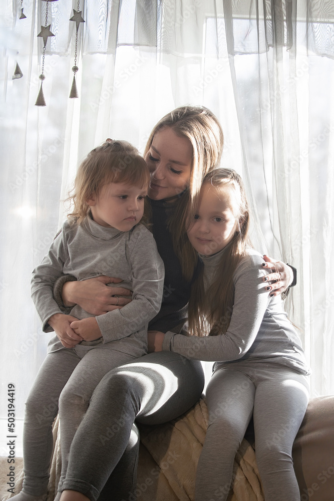 Portrait of a beautiful young woman with two little daughters at home against the background of the window. Family spend leisure time in cozy home environment Portrait of a beautiful young woman with two little daughters at home against the background of the window. Family spend leisure time in cozy home environment