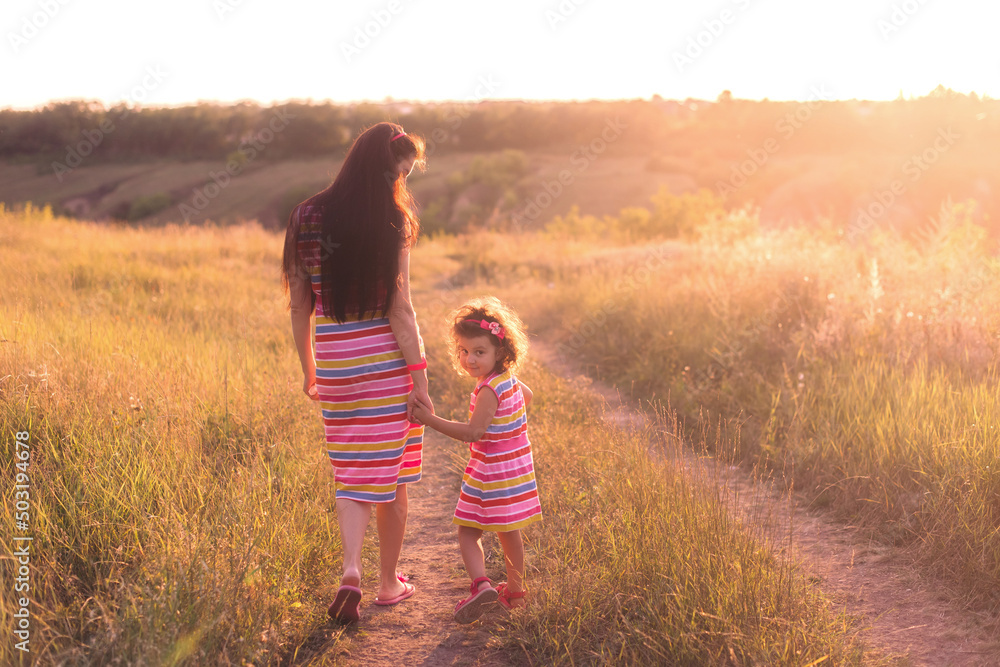 Happy mother with her daughter wearing in similar dresses walk holding hands on field at sunset. Mothers day .