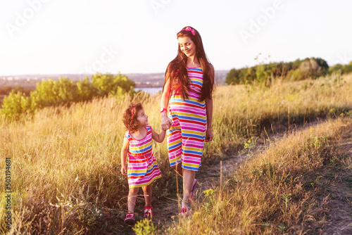 Obraz na plátně Happy pregnant mother with her daughter wearing in similar dresses walk holding hands on field at sunset