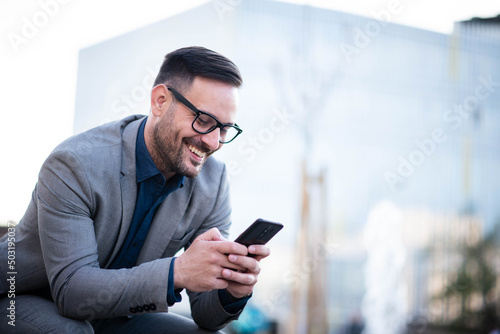 Business man using phone outdoors. Holding phone with both hands, texting and smiling
