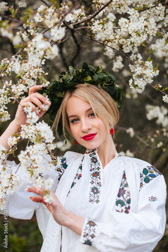 A Ukrainian woman in a white embroidered shirt and with a wreath on her head in a blooming garden in spring. Portrait of a Slavic girl.
