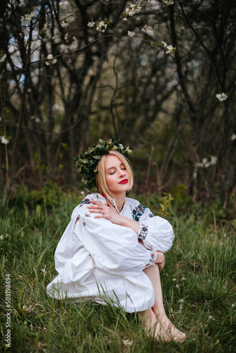 A Ukrainian woman in a white embroidered shirt and with a wreath on her head in a blooming garden in spring. Portrait of a Slavic girl.