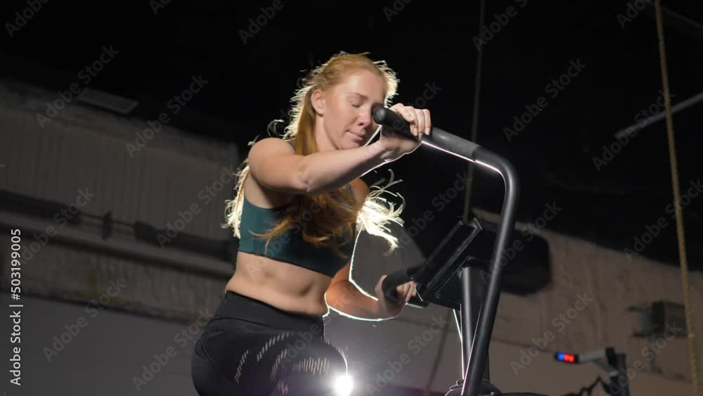 Young ginger-haired woman with high ponytail in sportswear trains actively developing muscles and strength in sports room at back spotlight Young ginger-haired woman with high ponytail in sportswear trains actively developing muscles and strength in sports room at back spotlight