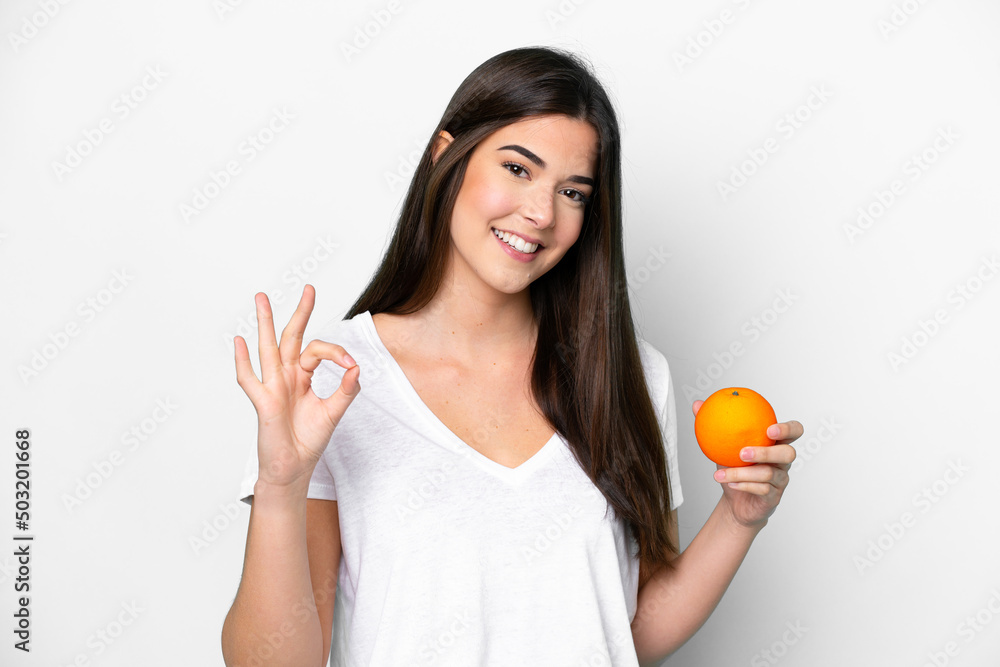 Young Brazilian woman holding an orange isolated on white background showing ok sign with fingers Young Brazilian woman holding an orange isolated on white background showing ok sign with fingers
