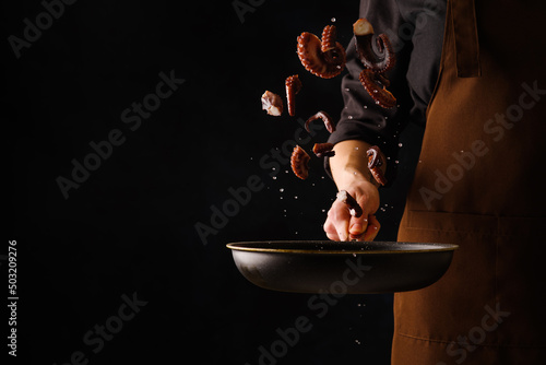 Fototapeta Naklejka Na Ścianę i Meble -  A professional chef cooks octopuses in a frying pan on a black background. Seafood in frozen flight. There is free space to insert. Seafood recipes. Restaurant, hotel, banquet, picnic.