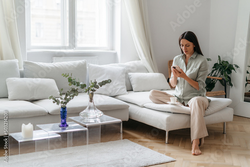 Happy pretty young woman in casual wear sitting on sofa at home and using smartphone while browsing internet