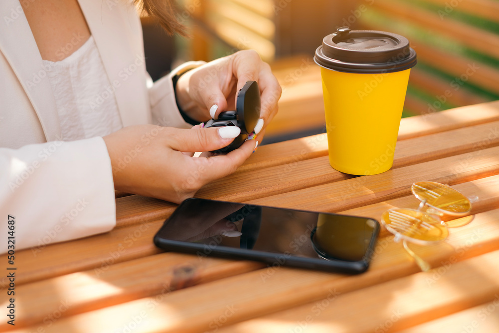 Close-up of a women's hands taking out of the box wireless small black earbuds. Girl at a table with a black mobile smartphone and a glass of coffee and sunglasses . White manicure