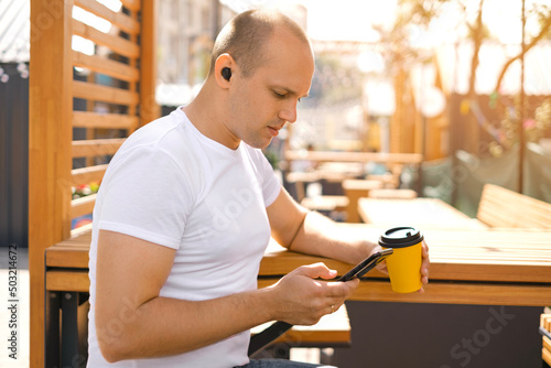 Man in a white T-shirt is sitting at table in a city street cafe with cup of coffee, listening to music and writing messages, chatting with friends. Modern technologies. Bluetooth earbuds. Free time