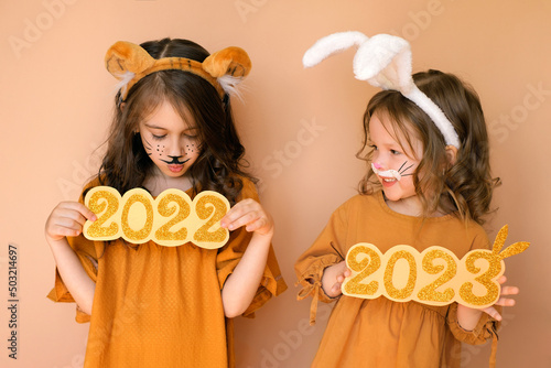 Outgoing year of Tiger and New Year of the Rabbit. Sad child with tiger makeup holds number 2022, and baby in guise of rabbit holds 2023. Painted face of people with aquagrim in image of animal.