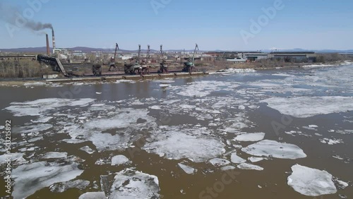 Ice Drift Occurs on the River next to the Port with Big Cranes and Factory, Mountains Ridge cover Forest, Aerial view photo