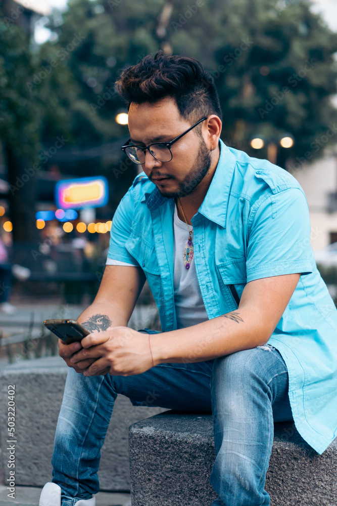 Fototapeta premium Latino man sitting on a bench wearing glasses, texting on his cell phone, in a city.