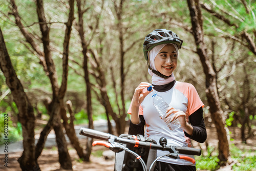 Sporty woman in hijab opening mineral water bottle during cycling break on outdoor background