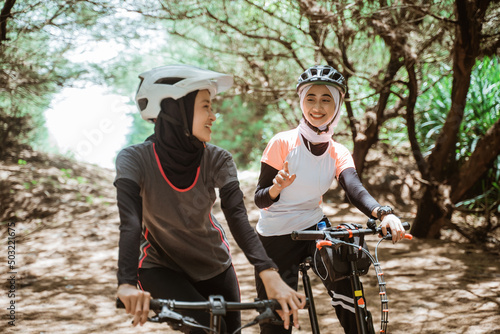 Two young Muslim women chatting while cycling outdoors with lots of trees