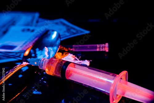 Syringe with cooked Heroin, white powder in a spoon and banknotes on a dark table with two-coloured lighting suggesting the forbidden and illegal. Concept of drug addiction, illegal trade. high view