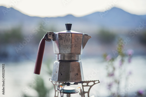 A retro coffee maker made of aluminum stands on a gas burner against the backdrop of mountain peaks on a hike. A tourist brews coffee in nature. 