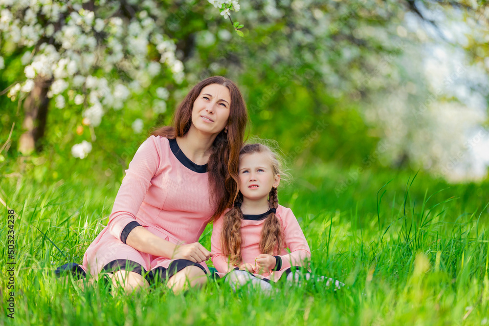 mother and daughter sit in the green grass against the backdrop of blooming apple trees mother and daughter sit in the green grass against the backdrop of blooming apple trees