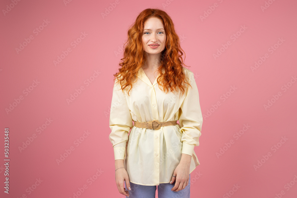 Picture of a beautiful redhead young woman with blue eyes looking at the camera dressed in yellow tunic standing on pink background.
