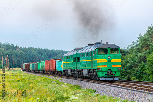 A powerful two-section green diesel locomotive pulls a long freight train to the railway station through a pine forest. Spring weather. Evening lighting.