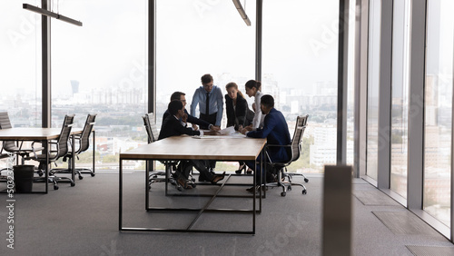 Diverse millennial business team meeting at modern office conference table, negotiating on project, investment, discussing work issues in contemporary interior with glass wall, big panoramic window