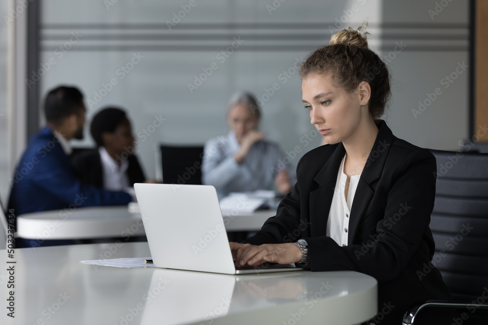 Focused pretty young employee woman using laptop computer in co-working space. Business professional lady working on online project, typing. Diverse team meeting in background Focused pretty young employee woman using laptop computer in co-working space. Business professional lady working on online project, typing. Diverse team meeting in background