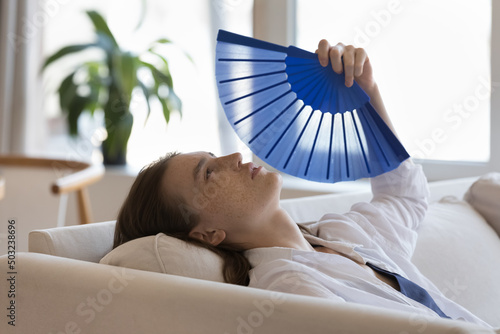 Tired sick millennial girl suffering from heat, hypoxia at home without conditioner, using paper fan, blowing cool air hot air, feeling unwell due to summer weather, humidity, high air temperature photo