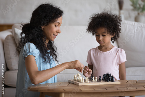 Engaged African mother teaching focused clever preschool daughter kid to play chess. Mom and girl thinking over game strategy at chessboard, planning next move. Family hobby concept
