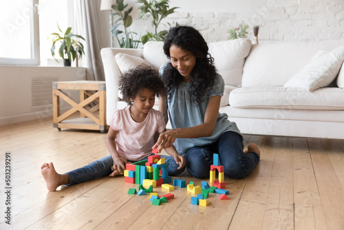 Positive daycare sitter teaching preschooler girl to build towers from toy blocks. Caring African American mother playing with daughter kid on warm heating floor, constructing model