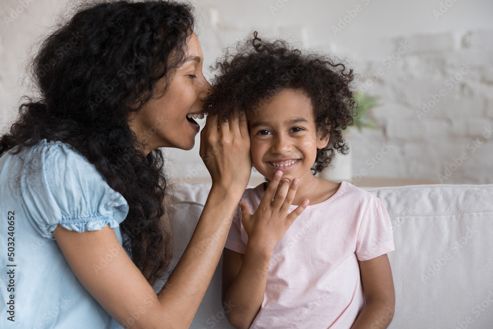 Cheerful Black mom telling secret to excited happy little girl, whispering in ear. African kid looking at camera, laughing. Mother and daughter enjoying friendship, trust, having fun at home Cheerful Black mom telling secret to excited happy little girl, whispering in ear. African kid looking at camera, laughing. Mother and daughter enjoying friendship, trust, having fun at home