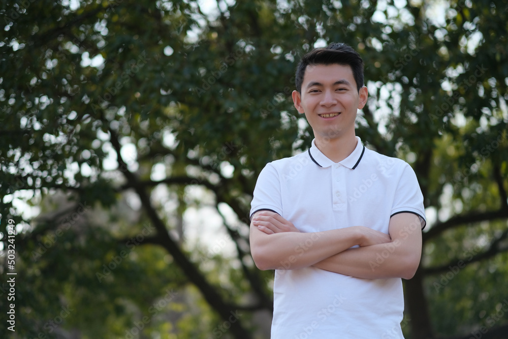 Chinese young man in white short sleeve polo shirt, cross his arms, smile and looking at camera in green park Chinese young man in white short sleeve polo shirt, cross his arms, smile and looking at camera in green park