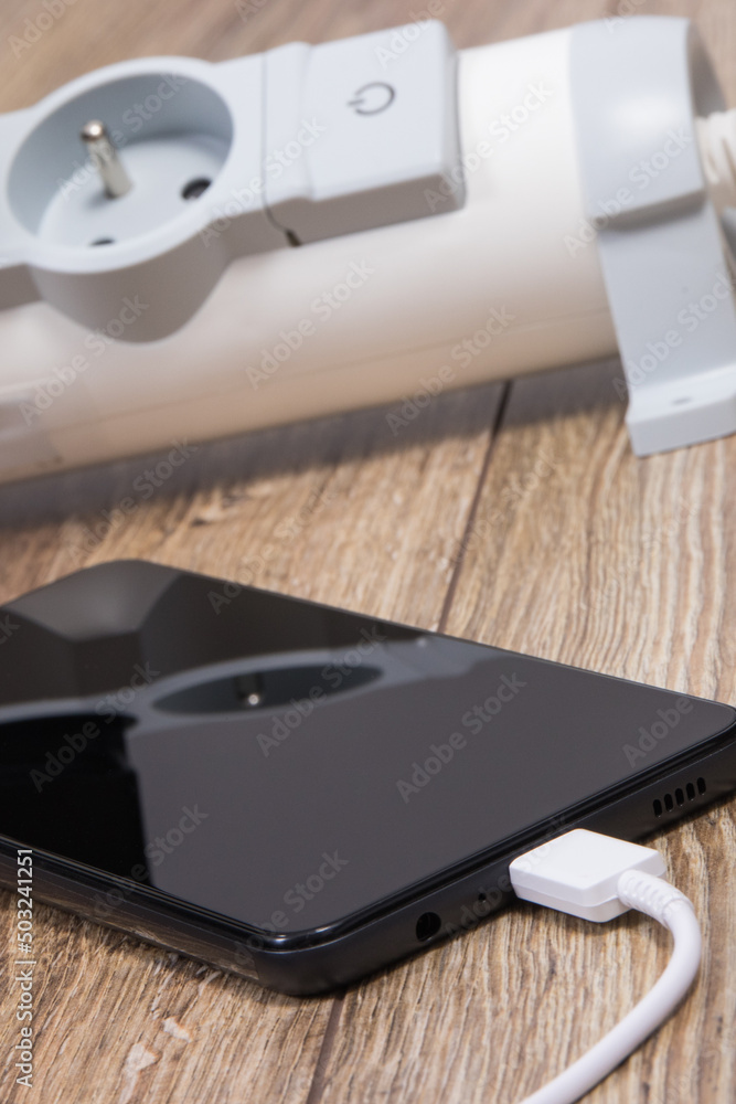 Black smartphone, connected white cable of charger and power strip in background. Telephone charging Black smartphone, connected white cable of charger and power strip in background. Telephone charging