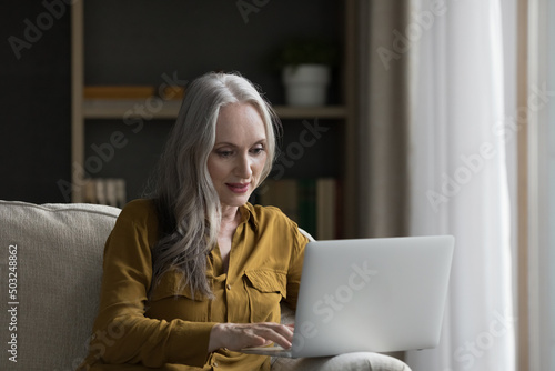 Focused pretty mature grey haired woman using telehealth app on laptop, consulting doctor online, typing, making video call, sitting on couch. Senior lady chatting on internet, photo