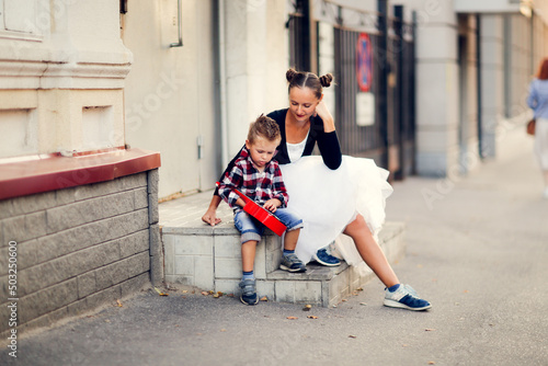 Cute european young mother with her son playing the red ukulele on the porch on the sidewalk. Walking around the city, learning to play musical instruments