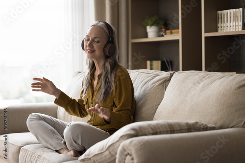 Cheerful carefree pretty mature woman wearing wireless headphones, listening to music, sitting on sofa in lotus pose, meditating, making dance hands, smiling, singing songs with closed eyes