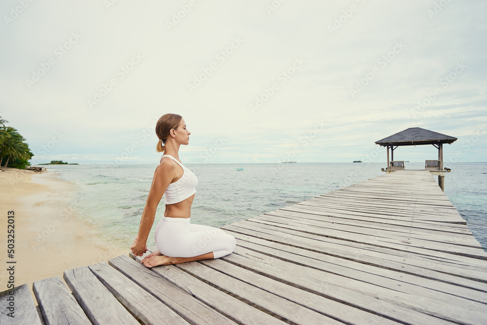 Yoga on the beach. Young woman stretching on wooden pier with sea view.