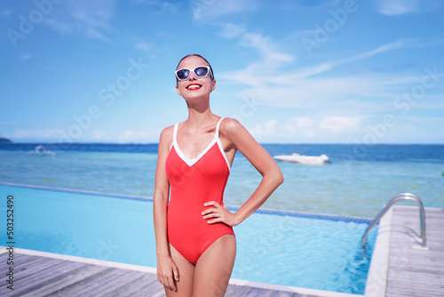 Summer vacation and fashion. Beautiful young woman in red swimsuit near swimming pool on tropical beach. © luengo_ua