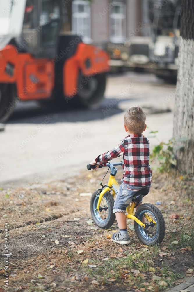cute european boy on balance bike watches how the road repair equipment works in the city, boys and construction equipment, boy looking at the skating rink cute european boy on balance bike watches how the road repair equipment works in the city, boys and construction equipment, boy looking at the skating rink