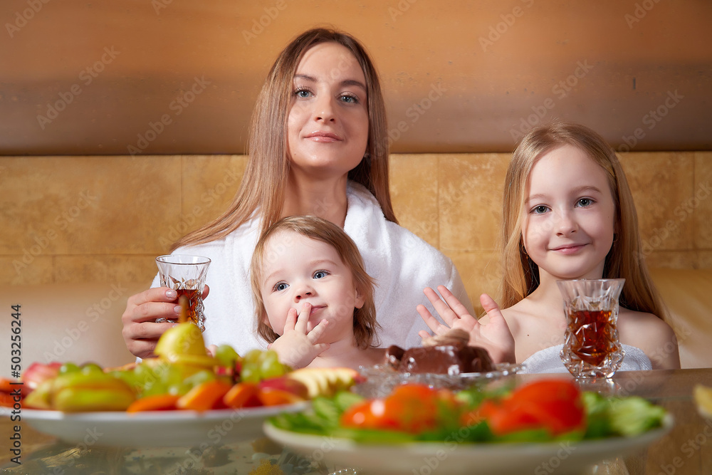 A little girl, female teenager and mother in a white bathrobe at a table with tee in a beautiful room in a Russian bath. The concept of family love, friendship and healthy holiday