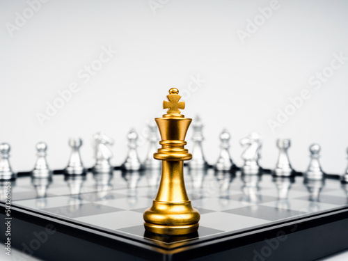 Fotografia The Golden king chess piece standing on chessboard corner in front of many silver chess pieces on white background