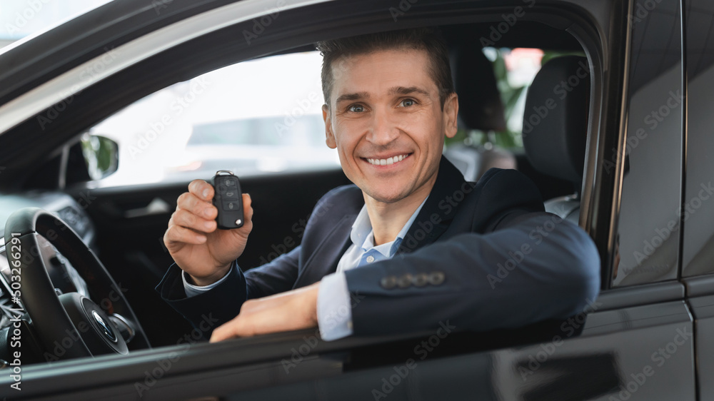 Happy insurance agent sitting inside new car, showing key from auto window, smiling at camera in automobile dealership