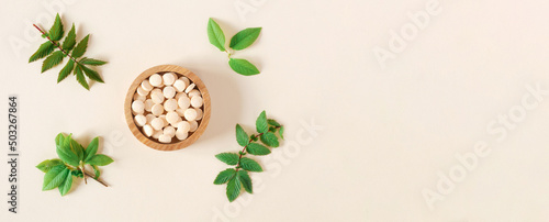 Directly Above Shot Of Pills and green leaves. Tablets in wooden bowl on beige neutral colors background. Homeopathic medicine. Organic medical capsules with herbal plant. Table top view. Copy space
