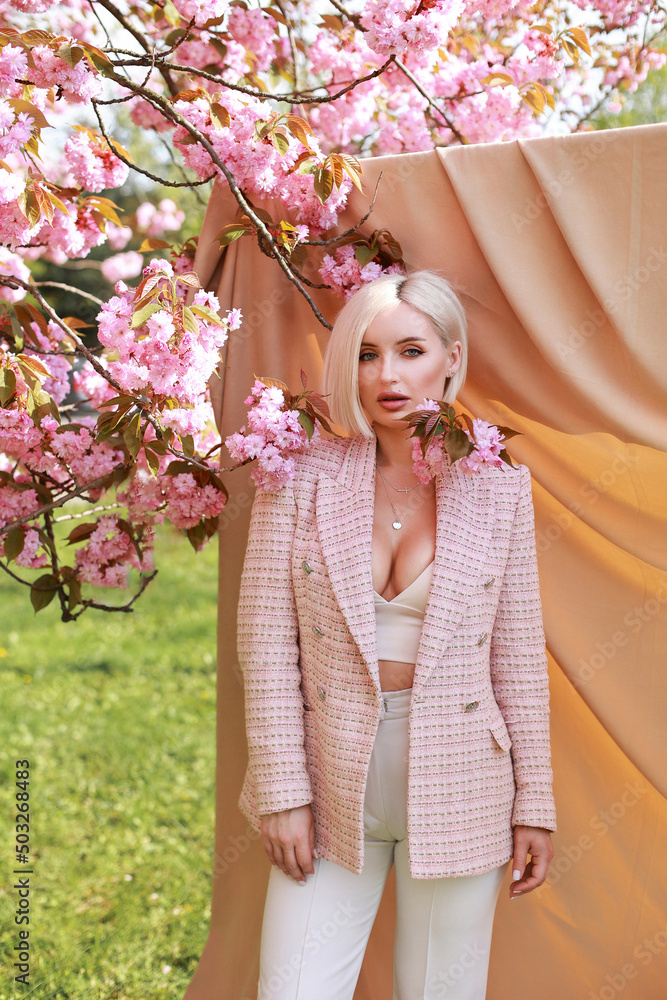 Fototapeta premium Gorgeous woman with short white hair on a beige background in the park near the sakura, which bloomed in spring with pink flowers.