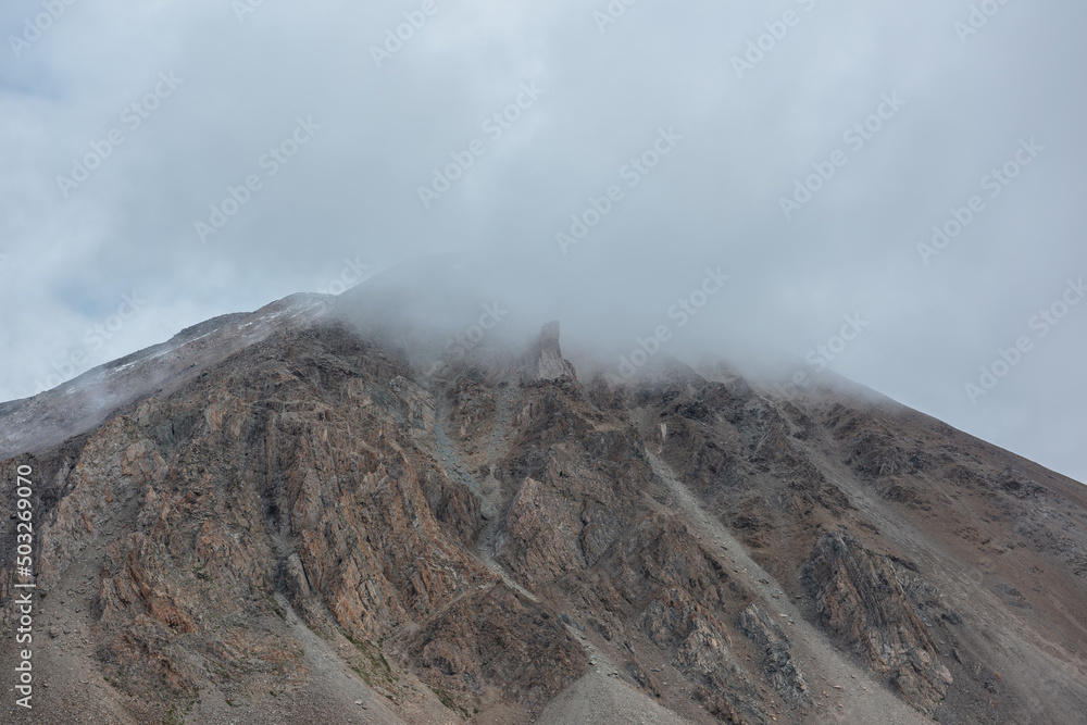 Dramatic mountain landscape with sharp rocks in rainy low clouds. Beautiful alpine scenery with large rocky mountain with snow top in low gray cloudy sky. Gloomy gray rainy weather in high mountains.